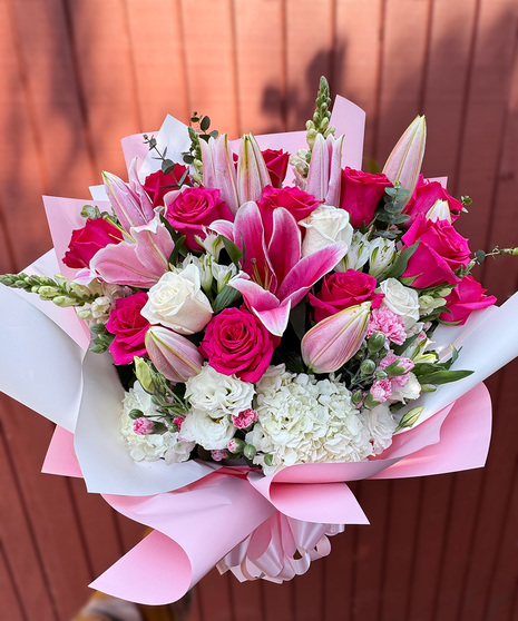 Blossom Glow Bouquet featuring fuchsia roses, white hydrangea, pink lilies, and pink stargazer lilies wrapped in pink decorative paper.